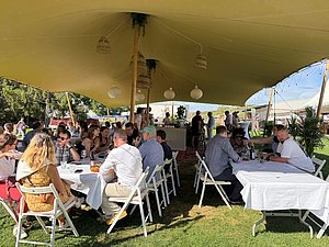 Menschen sitzen gemütlich bei Tisch und essen unter einem schattigen Zeltdach.